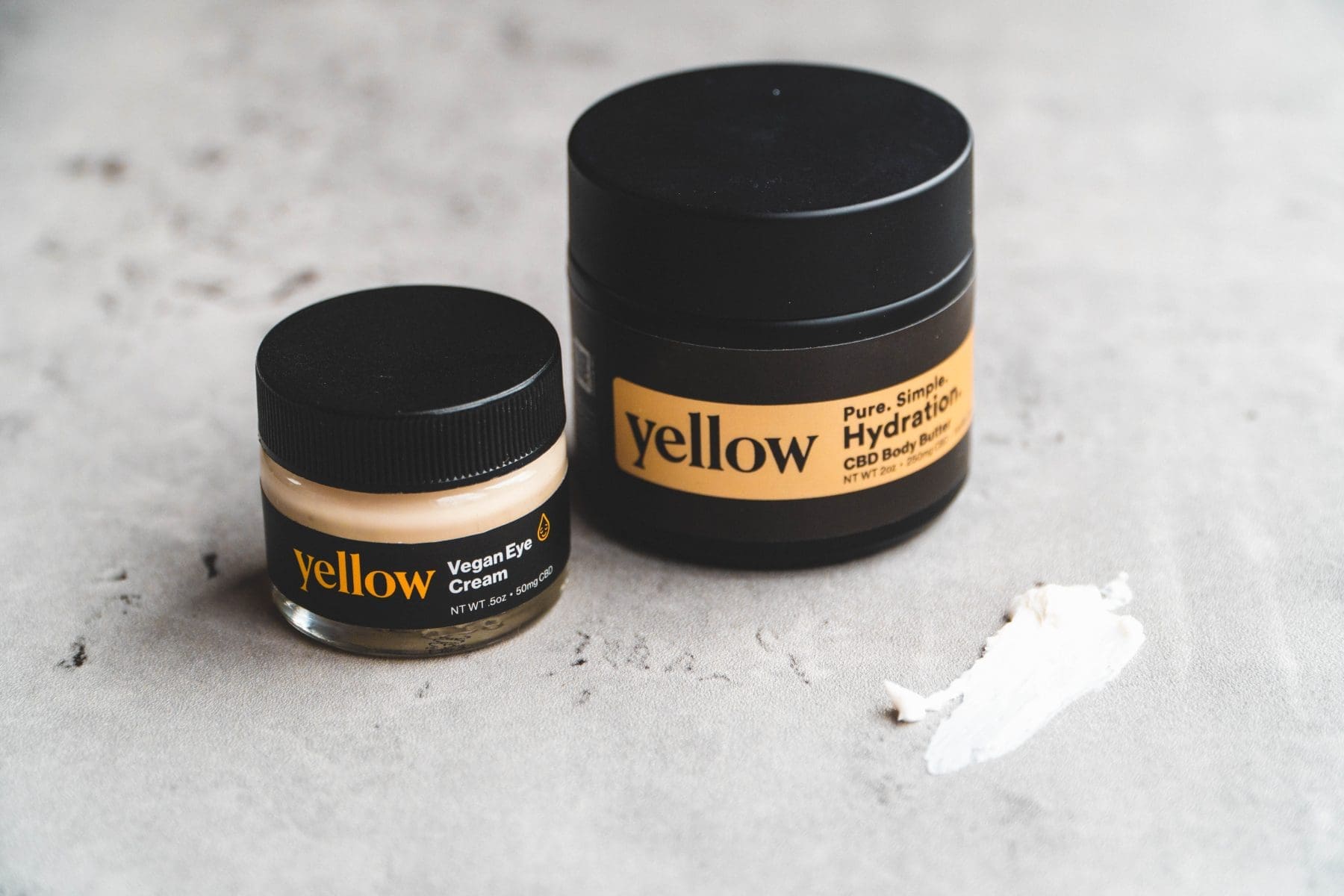 A jar and a smaller container of skincare products on a textured surface. The larger jar is labeled 'Hydration' with a black lid and a yellow band, while the smaller one is labeled 'Vegan Eye Cream.' A small amount of cream is smeared beside them.